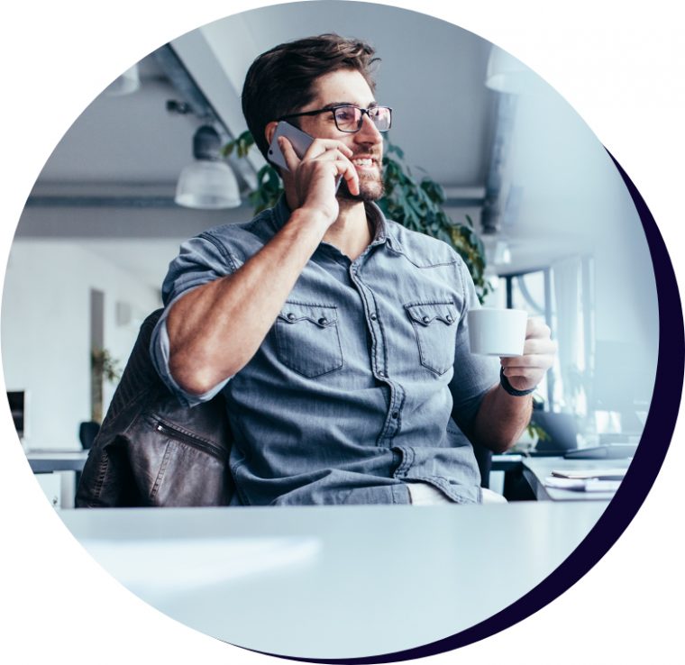 Man in collared shirt drinking a coffee at his office desk while on a mobile call